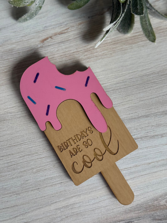 Ice cream popsicle-shaped laser-cut gift card holder with pink top and 'Birthdays are so cool' text on a wooden surface.