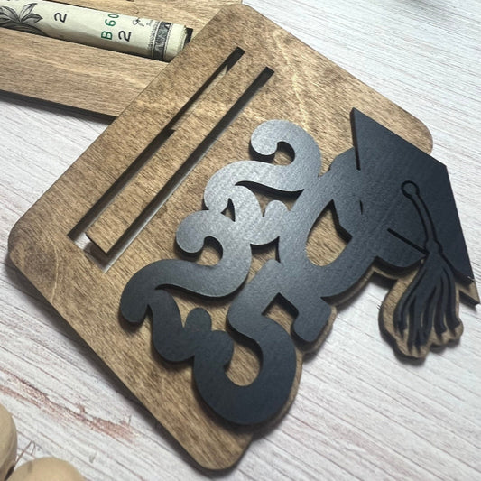 Laser cut cash holder with cut-out design of a graduation cap and tassel on a light wooden surface.
