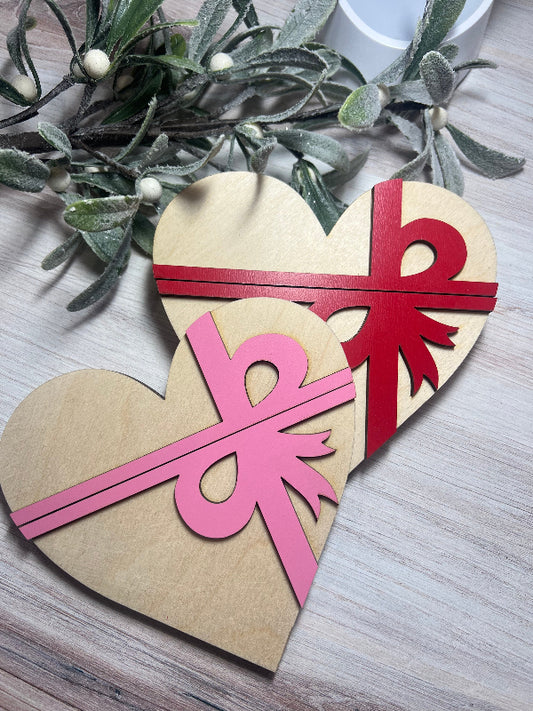 Heart-shaped wooden cutout with pink and red ribbons on a light wooden surface.