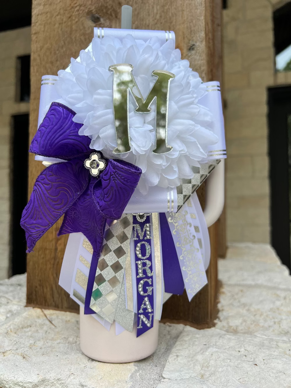 Decorative mini mum with a purple bow, white flowers, and a letter 'M' on a wooden surface.