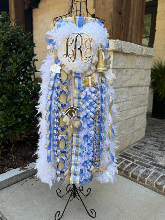 Decorative mum on a stand with blue, white, and gold ribbons and a monogram. Senior mum, The Woodlands Christian Academy