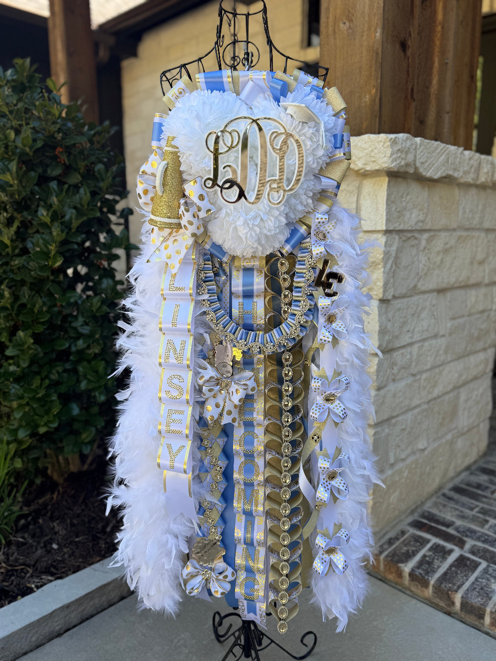 Decorative homecoming mum with ribbons and flowers on a stand outdoors. Columbia blue, white and gold.  Triple mum, Lake Creek High School.
