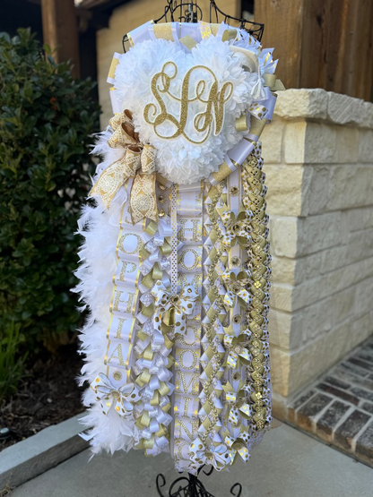 Decorative homecoming mum with white and gold ribbons on a stand against a stone wall. Triple mum, senior, Lake Creek High School.