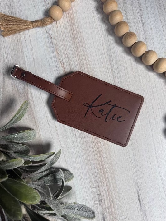 Brown leather luggage tag with 'Katie' engraving on a light wooden surface with decorative elements.