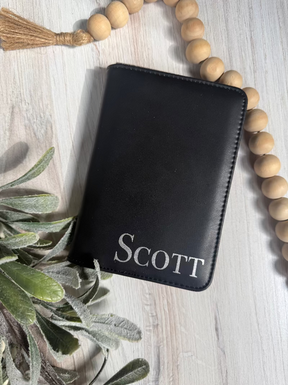 Black leather passport wallet with 'SCOTT' engraving on a light wooden surface with decorative elements.
