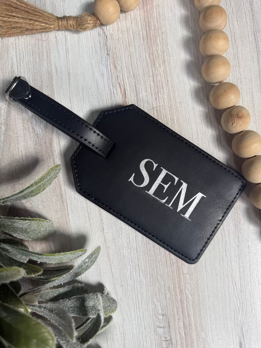 Black leather luggage tag with 'SEM' initials on a light wooden surface with decorative elements.