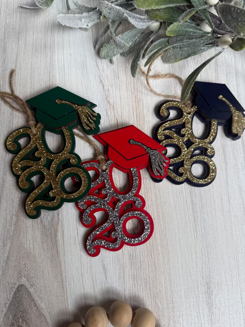 Decorative ornaments with graduation caps and '2026' text on a wooden surface with greenery.