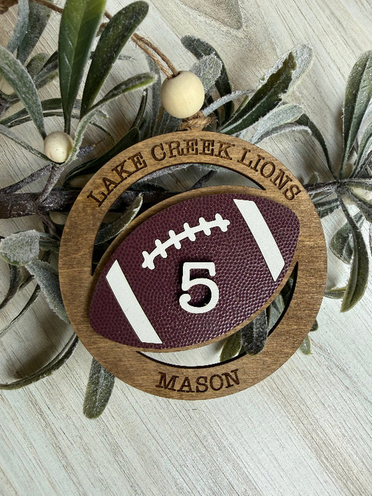 Decorative ornament with football design and 'Lake Creek Lions' text, placed on a textured surface with leaves.