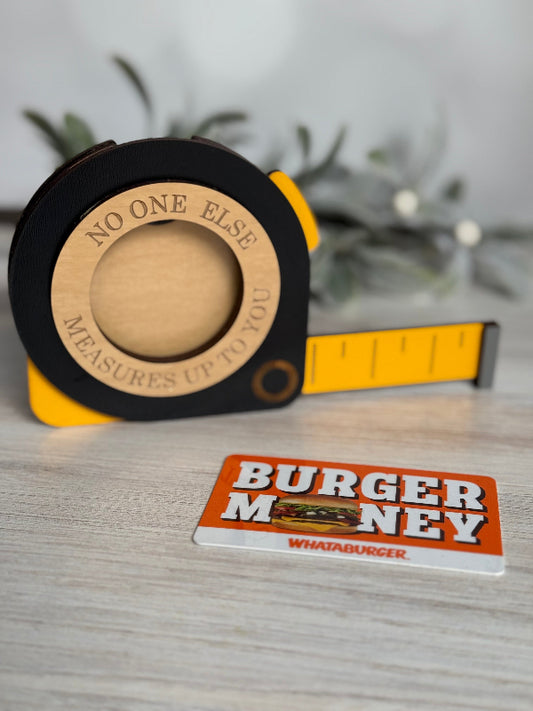Wooden measuring tape with a yellow band and a 'Burger Money' card on a wooden surface.